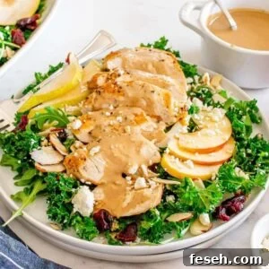A plate of autumn salad with chicken, featuring kale, apple slices, pear slices, almonds, cranberries, and crumbled cheese topped with grilled chicken and a creamy dressing. A bowl of dressing, an apple, and a pear are beside the plate.