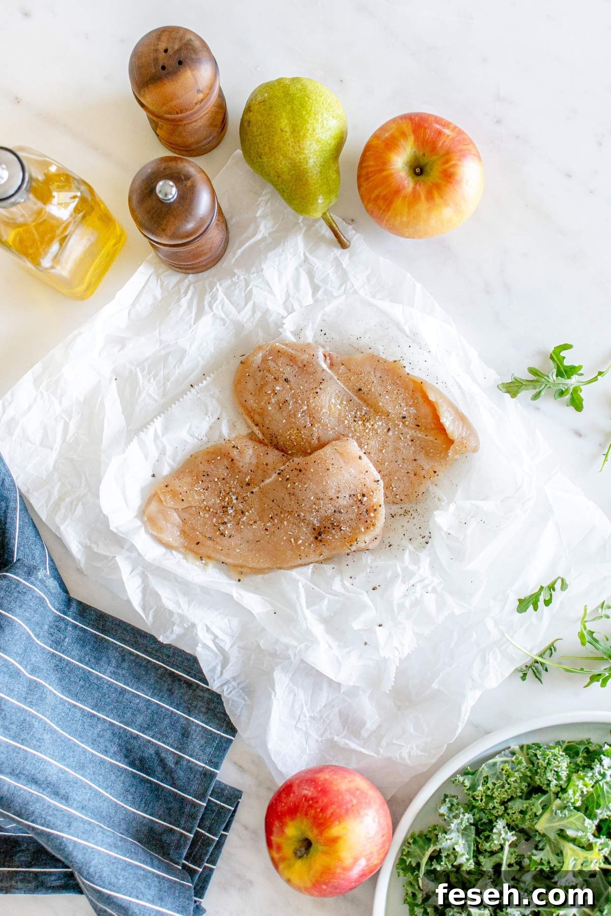 Raw, seasoned chicken breasts rest on parchment paper amidst a pear, apples, salt and pepper shakers, olive oil, a striped cloth, and a bowl of kale—everything you need for an autumn salad with chicken on a marble surface.