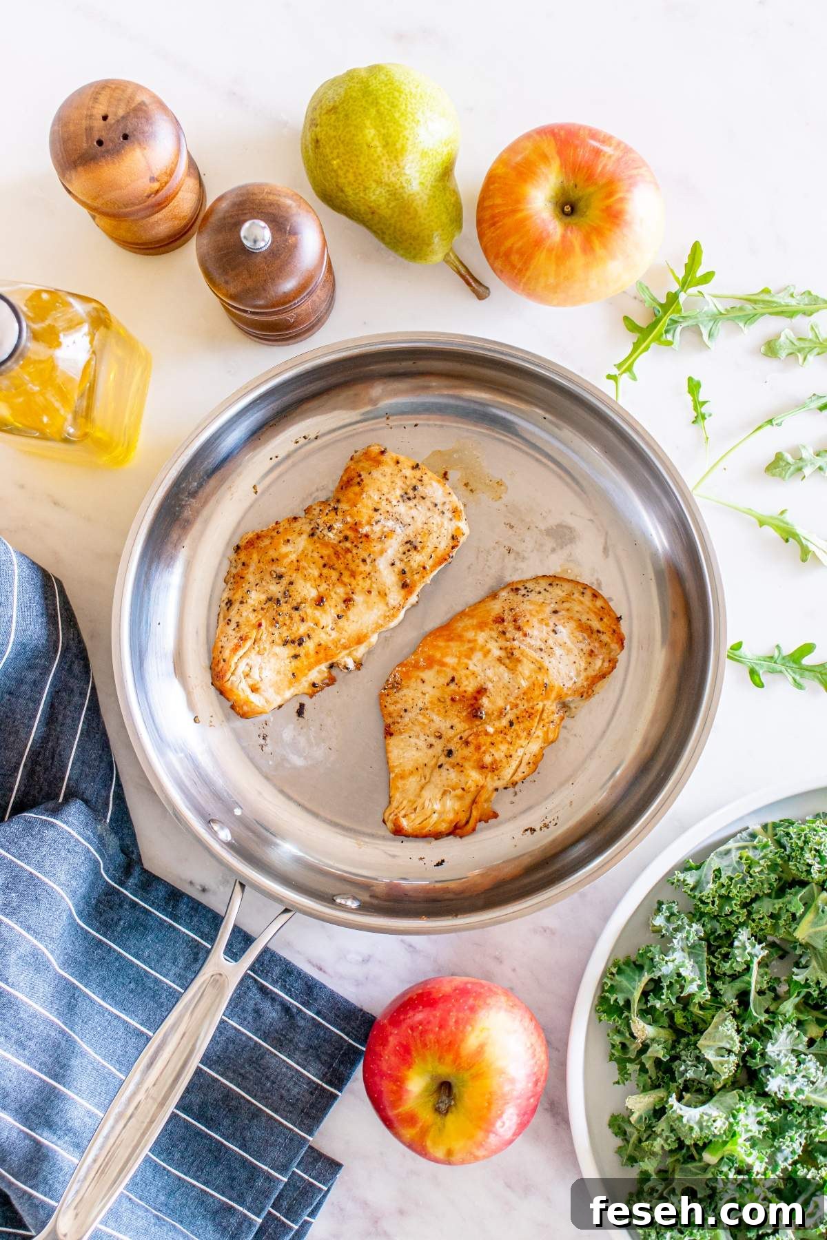 Two cooked chicken breasts in a stainless steel skillet, surrounded by an apple, a pear, kale salad, olive oil, salt and pepper shakers, and a striped cloth napkin on a white marble surface—perfect for an autumn salad with chicken.