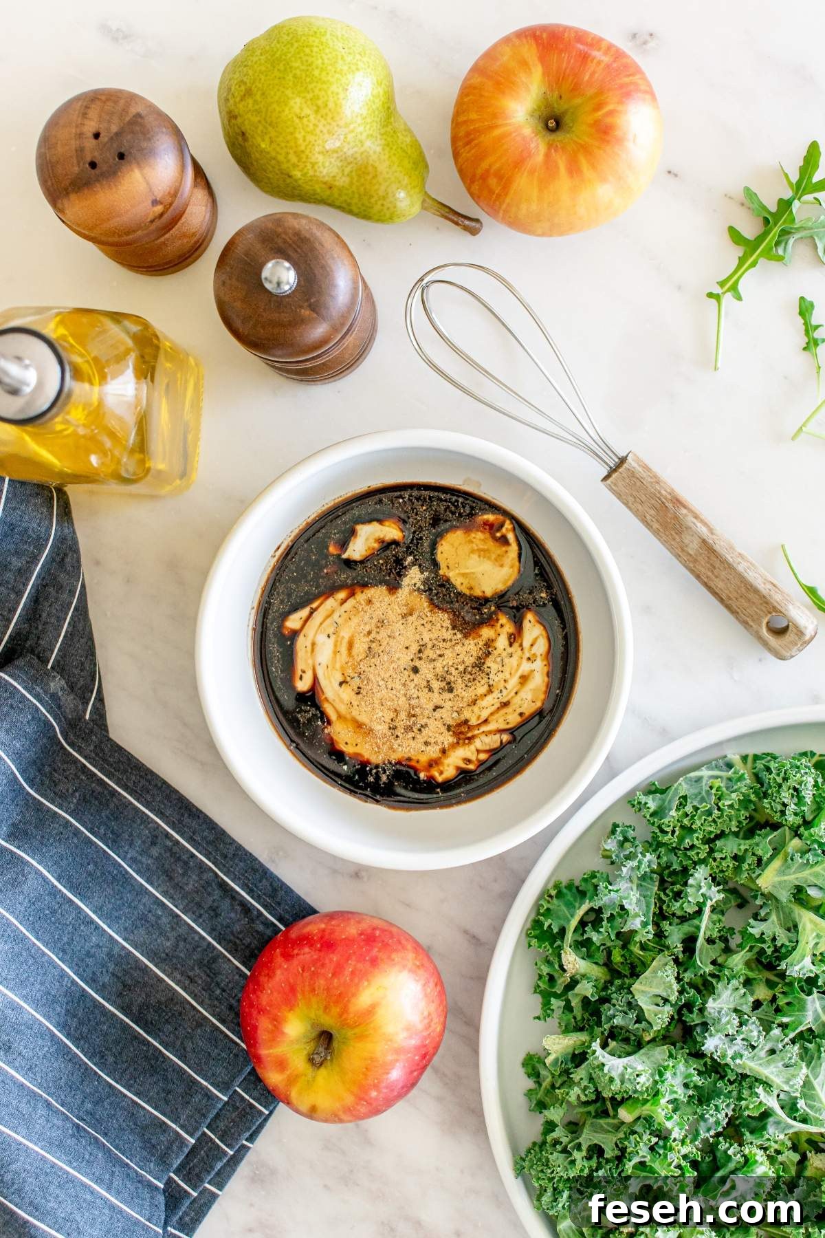 A bowl of balsamic vinaigrette, a whisk, apples, a pear, olive oil, salt and pepper grinders, a striped cloth, and a plate of kale on a white countertop set the stage for preparing an autumn salad with chicken.