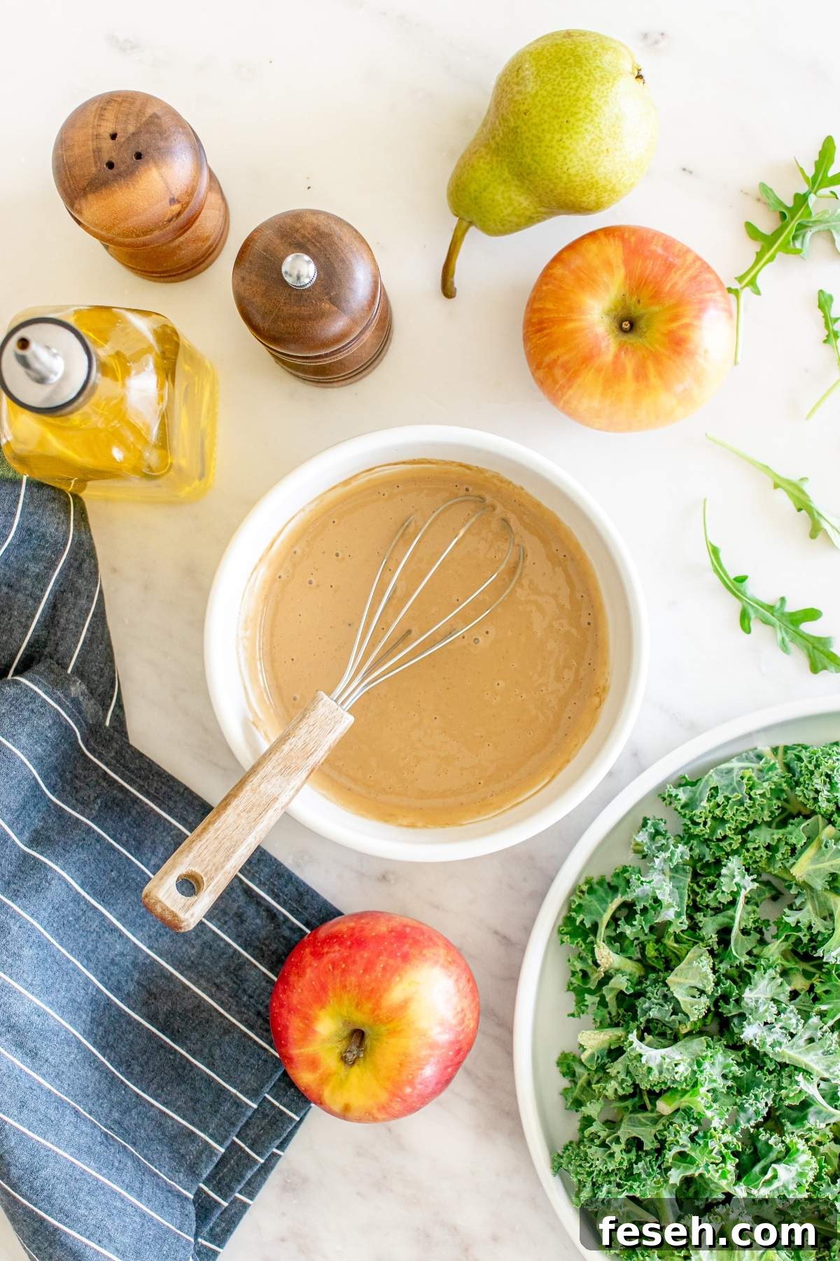 Top view of a whisk in a bowl of brown sauce surrounded by apples, pear, kale, olive oil bottle, salt, pepper shakers, a striped cloth, and arugula on a white surface—perfect for preparing an autumn salad with chicken.