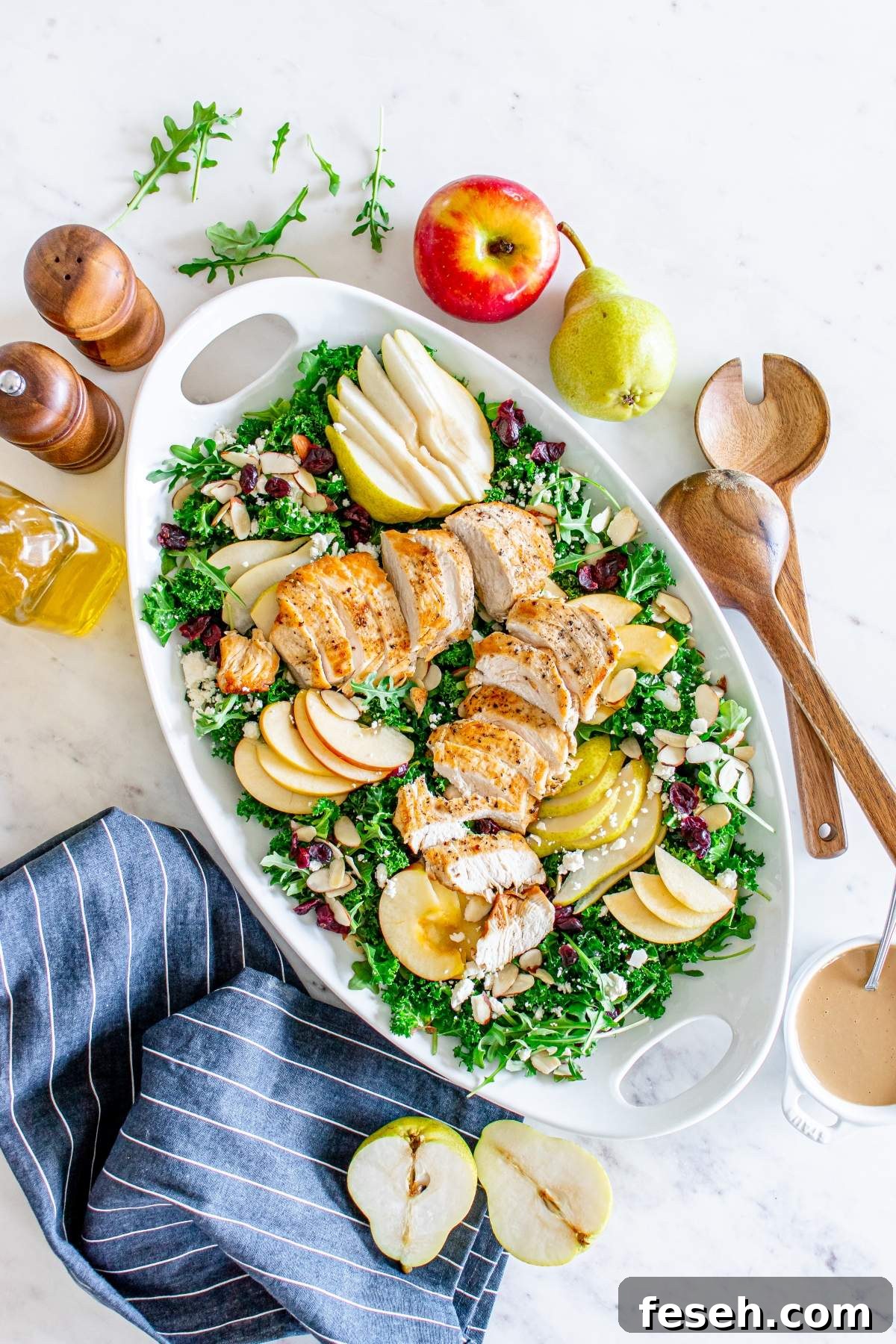 An autumn salad with chicken features mixed greens, sliced grilled chicken, apple and pear slices, nuts, cranberries, and cheese on a white platter. Accompanied by a striped napkin, salad utensils, fruits, dressing, and seasonings beside it.