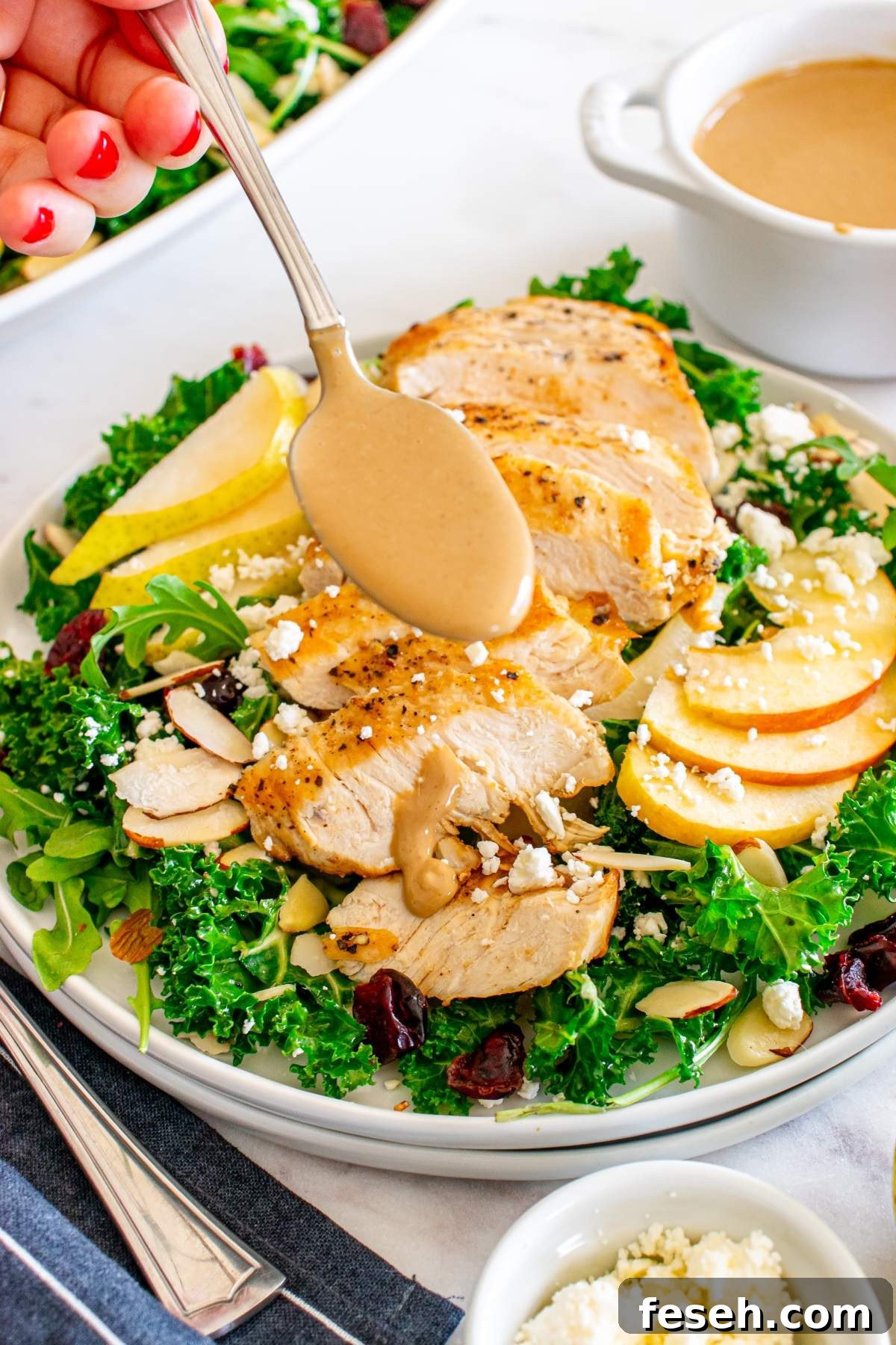A hand drizzles dressing over an autumn salad with chicken, featuring sliced chicken, kale, arugula, sliced apples, almonds, cranberries, and crumbled cheese on a white plate. A bowl of dressing is in the background.