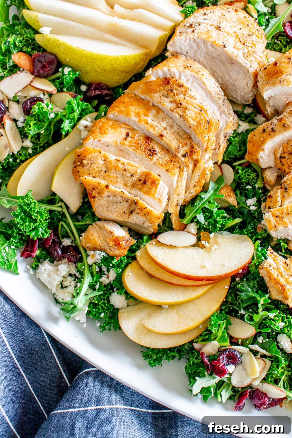 A fresh autumn salad with chicken, featuring sliced grilled chicken, kale, apple slices, pear slices, almonds, cranberries, and crumbled cheese on a white plate.