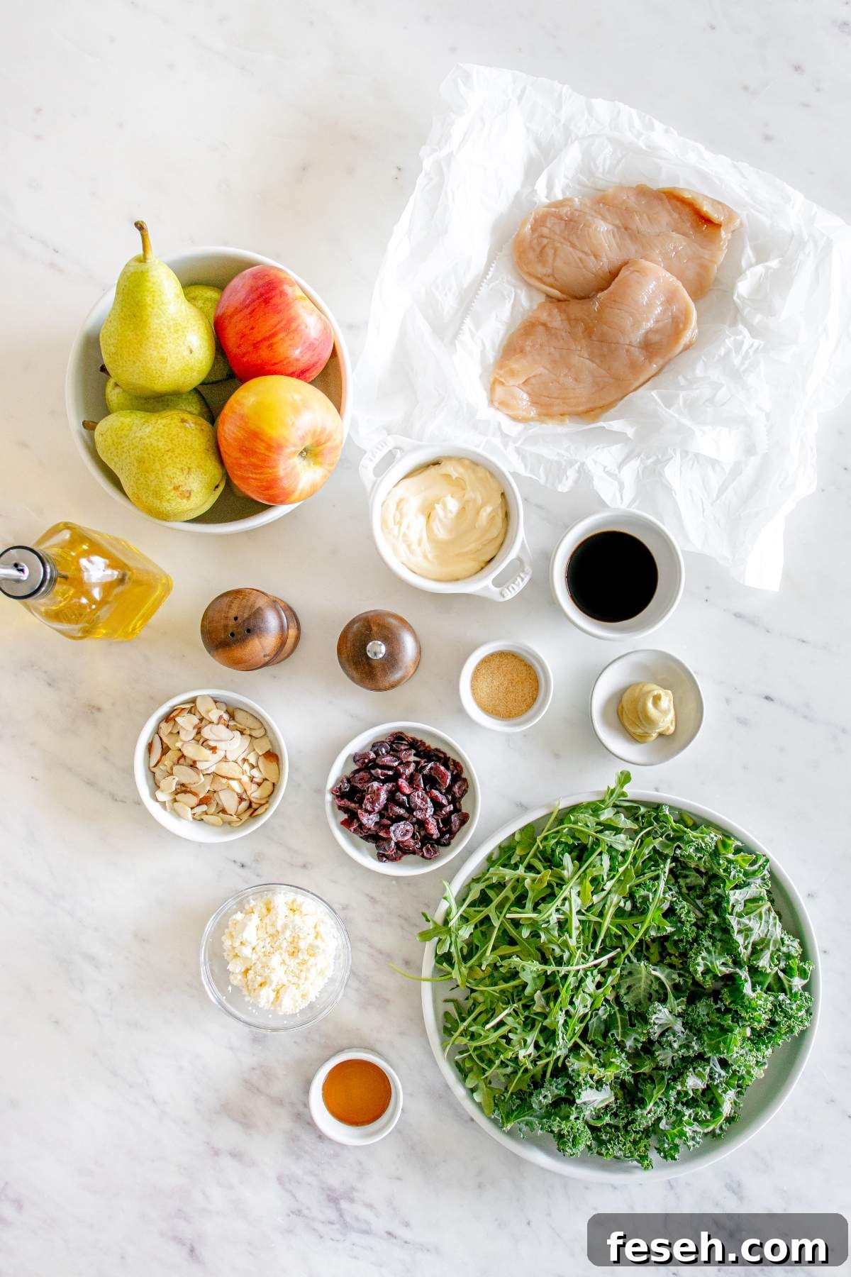 A marble surface displays ingredients for an autumn harvest salad with chicken, including pears, apples, chicken breast, olive oil, mayonnaise, soy sauce, mustard, brown sugar, sliced almonds, dried cranberries, feta cheese, arugula, and kale.