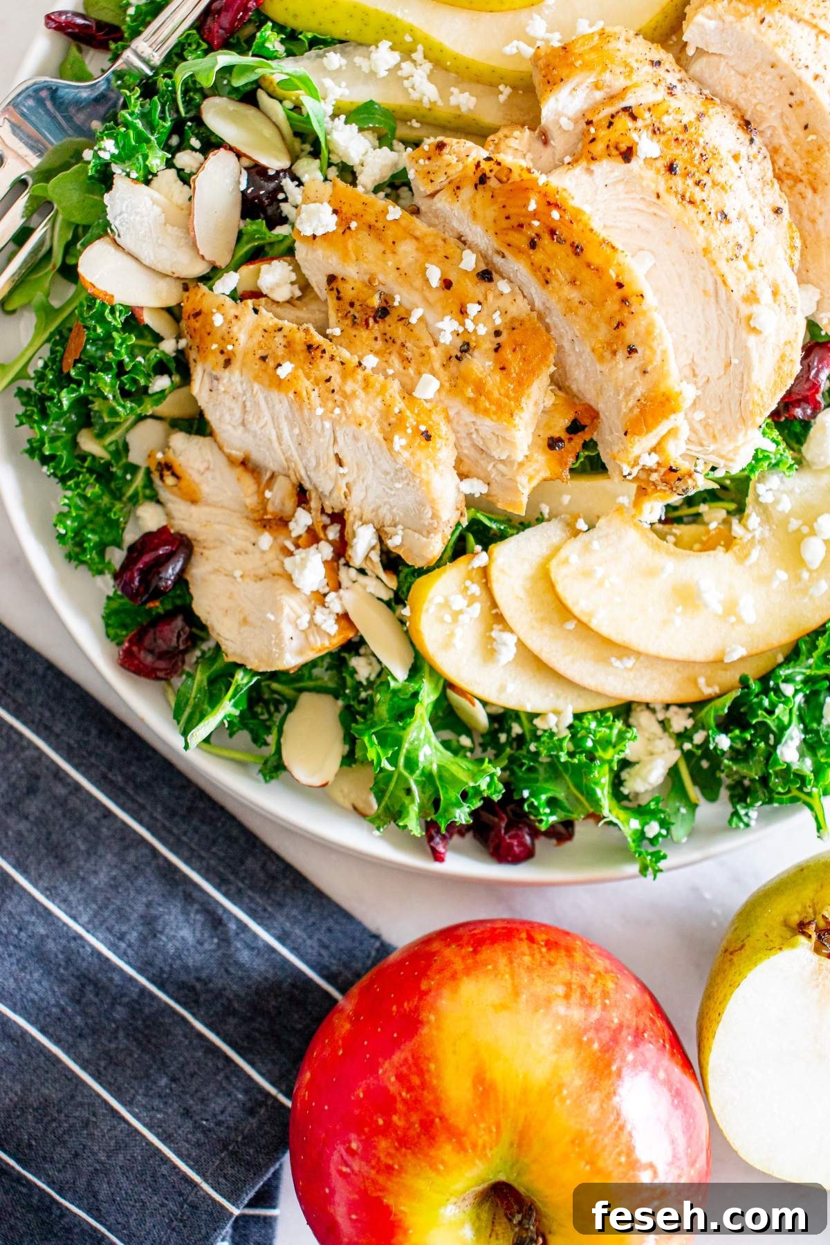 A plate of autumn crunch salad with chicken, featuring grilled chicken slices, apple slices, almonds, dried cranberries, and crumbled cheese, sits next to a red apple and a pear on a table with a striped napkin.