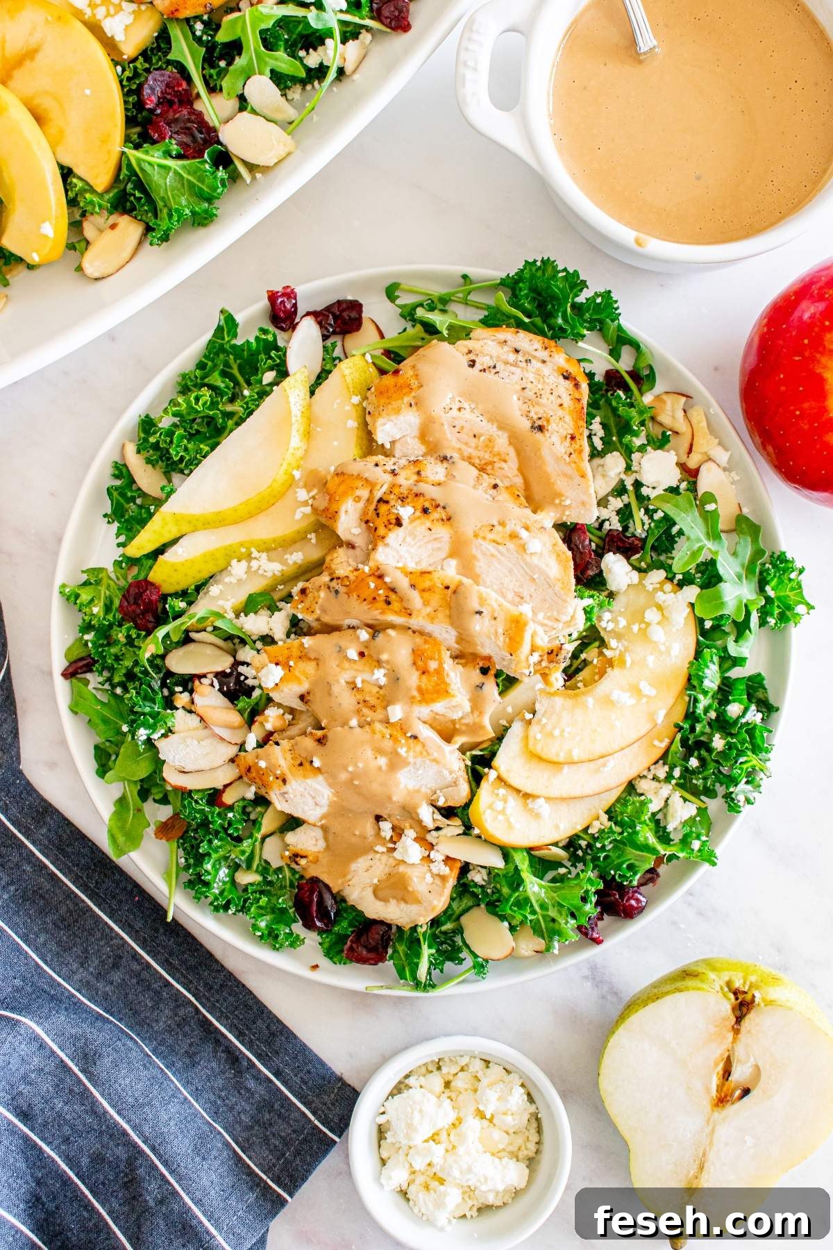 A crunchy autumn harvest salad with chicken slices, pear slices, almond slivers, and feta cheese is arranged on a white plate. A bowl of dressing and sliced pear sit beside it, with a striped cloth napkin nearby.
