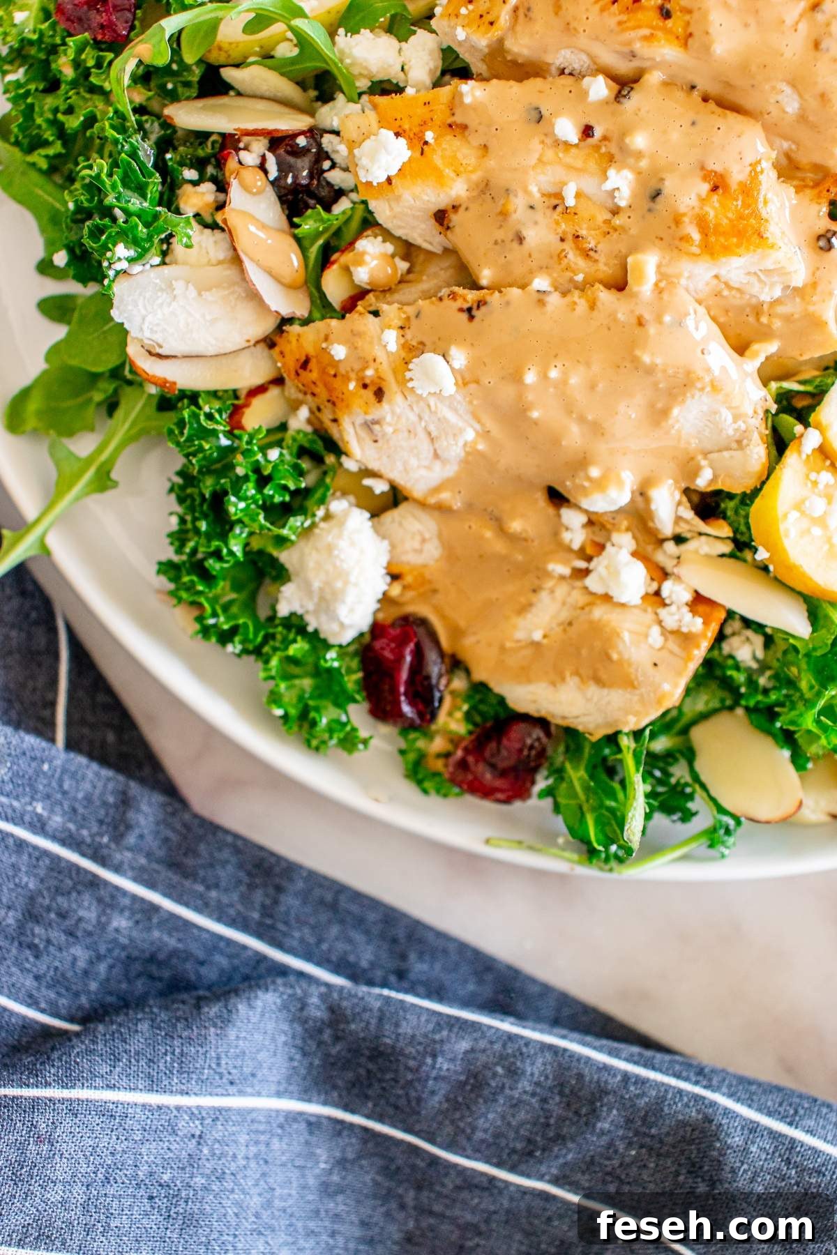 A plate of autumn apple pear kale arugula salad with chicken, creamy dressing, crumbled cheese, nuts, and dried cranberries. A striped fabric is partially visible in the foreground.