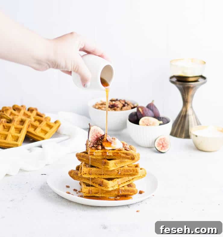 four orange pumpkin waffles stacked on top of each other with a fig, granola and butter with maple syrup poured over the top from a small white pitcher.