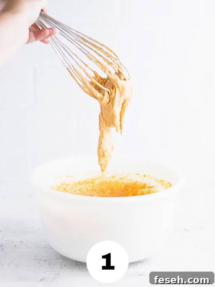 Whisk with thick, orange batter dripping off of it into a white bowl.