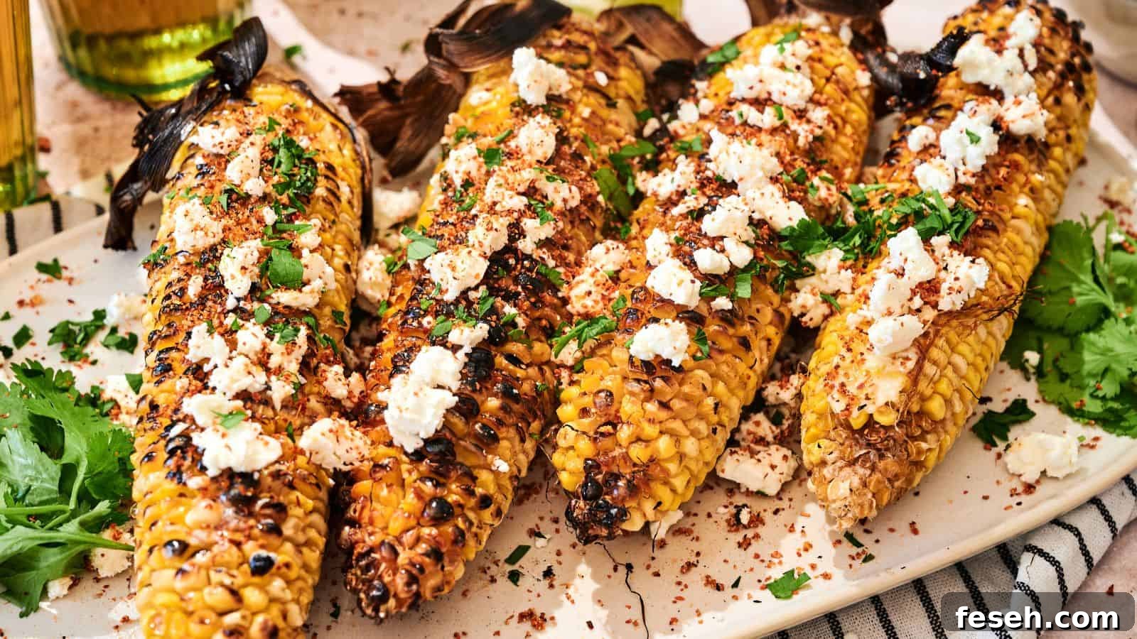 Four grilled corn cobs topped with crumbled cheese, chopped herbs, and spices are arranged on a white plate.