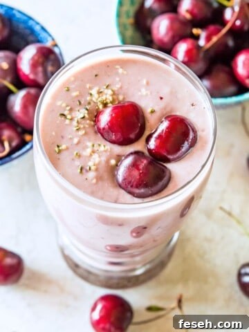A clear glass with a cherry smoothie in it, topped with fresh cherries and hemp hearts with fresh cherries around the glass.