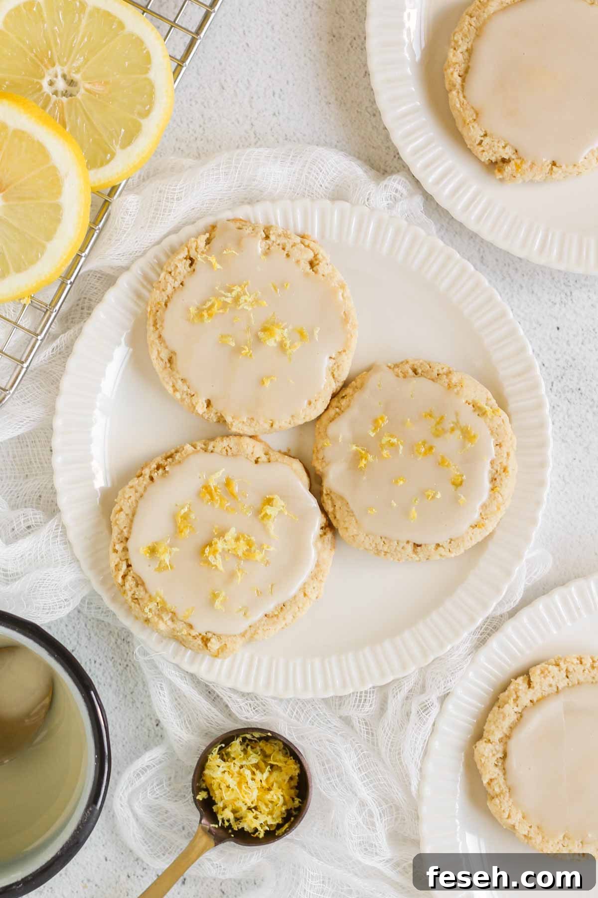 Three glazed lemon cookies sprinkled with lemon zest on a small plate with additional plates, lemons, and a bowl of glaze off to the sides.