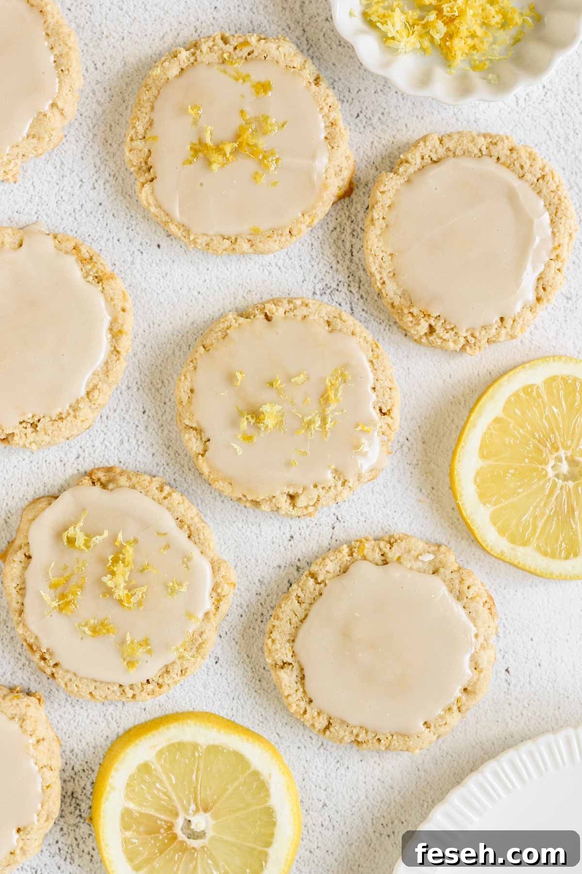 Glazed almond flour lemon cookies topped with fresh lemon zest, arranged on a light surface with sliced lemons and a small bowl of lemon zest nearby.