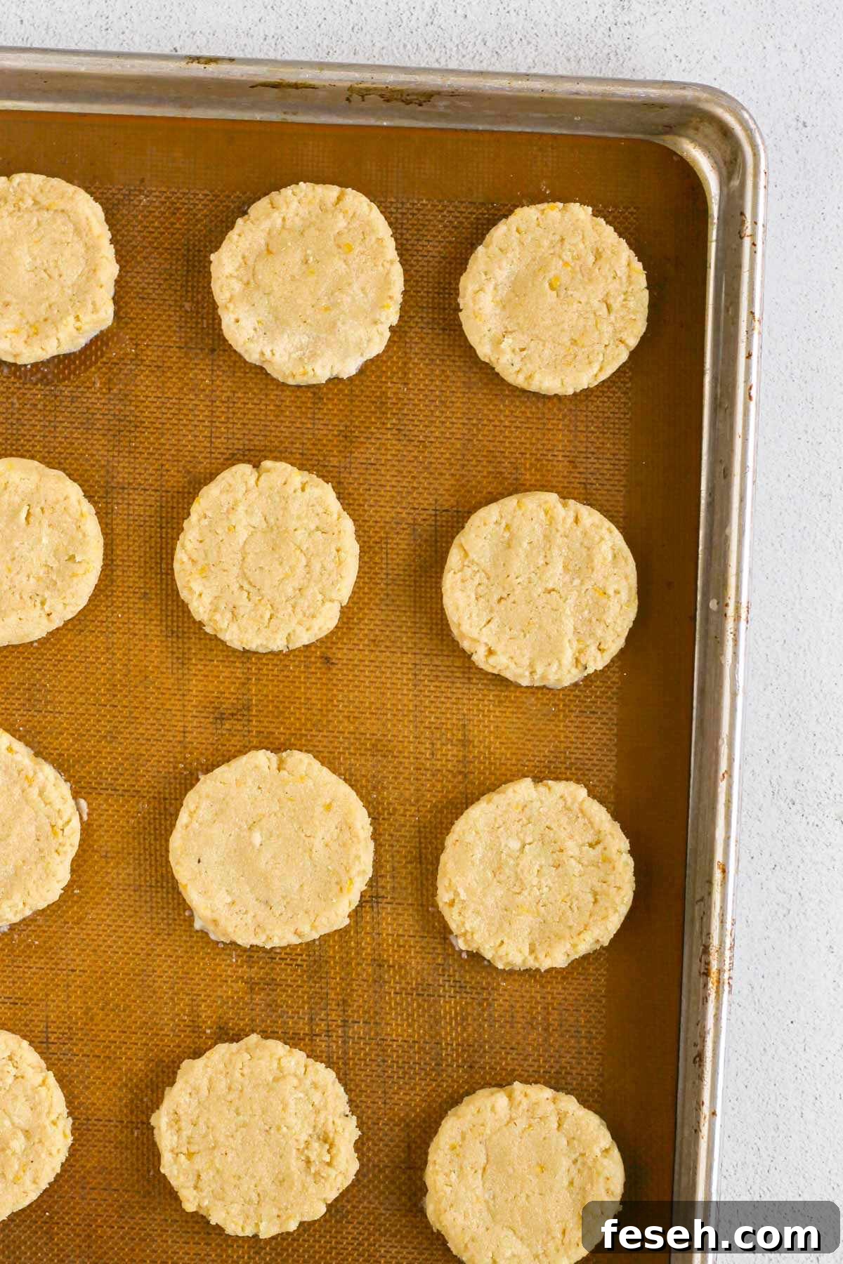 Almond flour cookie dough shaped into round cookies on a silicone mat lined baking sheet before being baked.