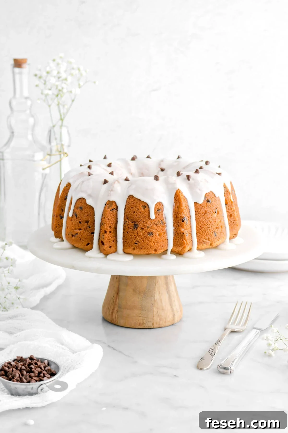 bundt cake on cake stand with icing and chocolate chips on top, a measuring cup of chocolate chips, a fork and knife, a white cheesecloth, and flowers beside.