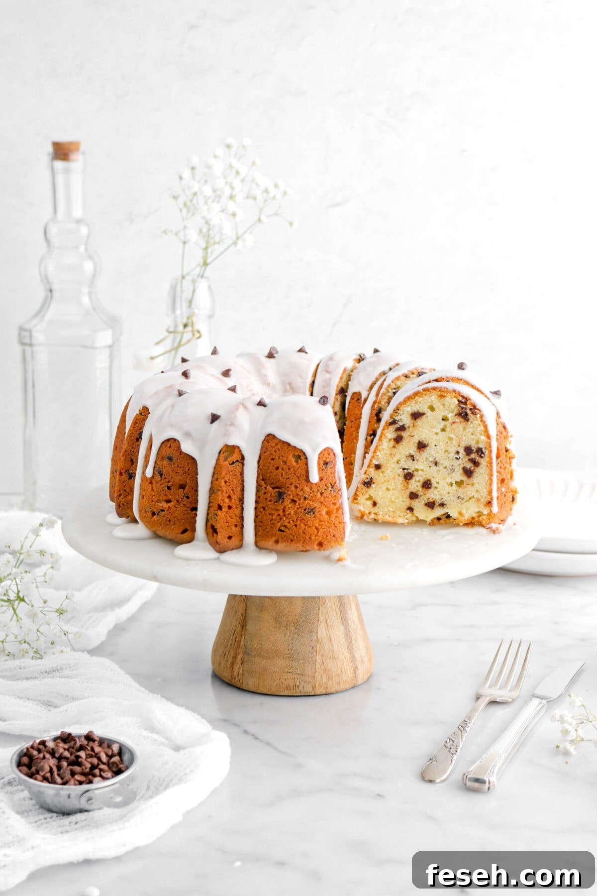 chocolate chip pound cake with five slices leaning against cake on a marble and wooden base cake stand, with fork and knife beside, a measuring up of chocolate chips, a white cheesecloth, and white flowers.