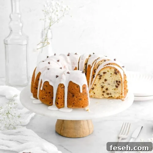 five slices of chocolate chip pound cake leaning against the rest of the cake on a cake stand, with stack of plates and empty glasses behind, a white cheesecloth beside, a measuring cup with mini chocolate chips, and a fork and knife beside on marble surface.