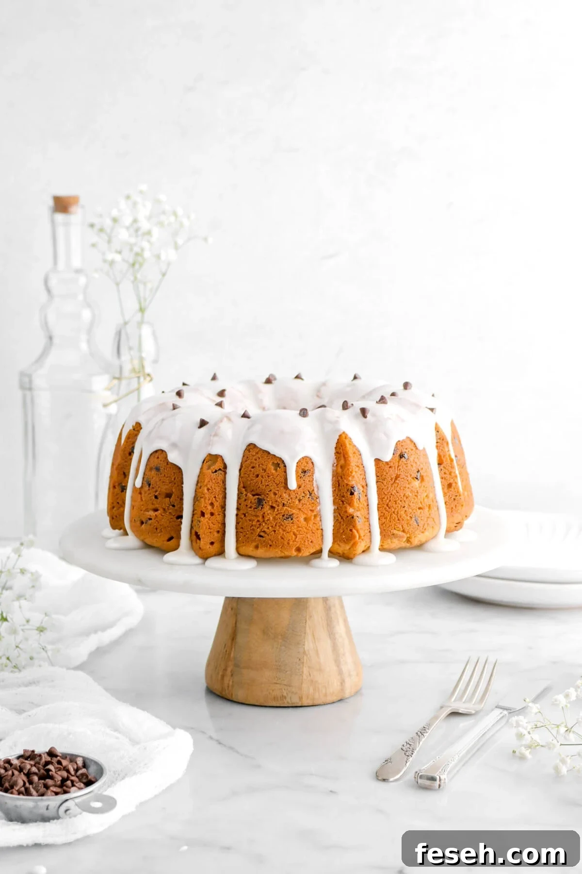 front shot of chocolate chip pound cake on marble cake stand with wooden base, a fork and knife beside, as well as flowers, and a measuring cup of mini chocolate chips on marble surface.