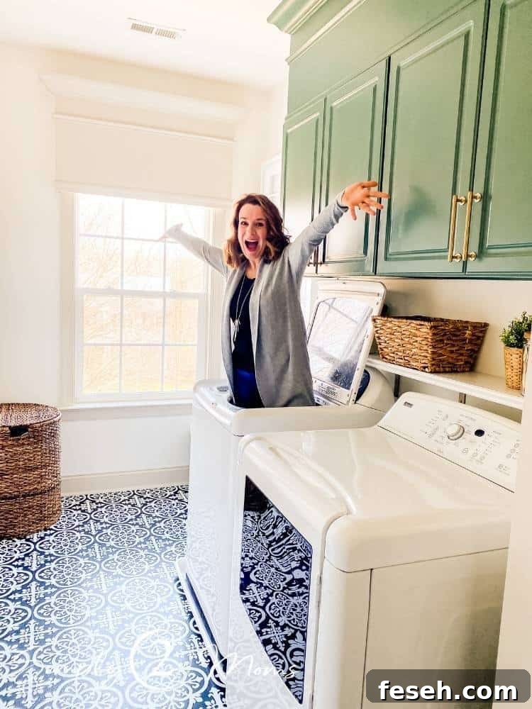 Woman standing inside a top-loading washing machine with arms raised and happy face showing off newly renovated French country farmhouse laundry room with painted green laminated cabinets, ceramic tile floors, and custom window valance.