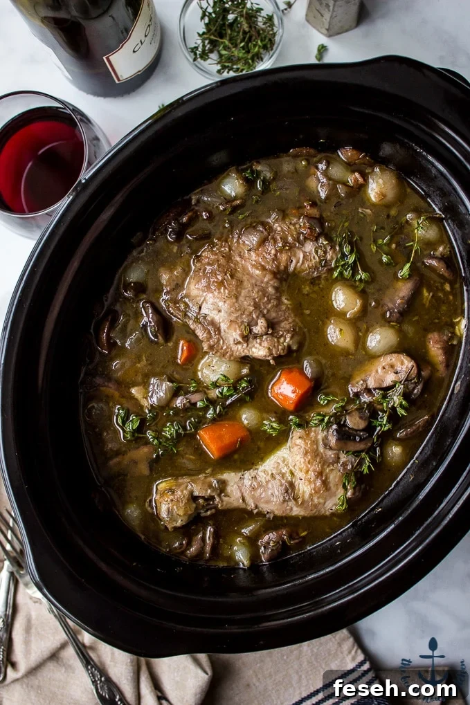 Overhead photo of Slow Cooker Coq Au Vin in a black slow cooker with a bottle of wine , glass of wine and salt and pepper shakers in background.