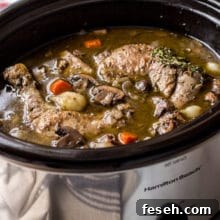 Coq Au Vin in a silver slow cooker with a bottle of wine and salt and pepper shakers in background.