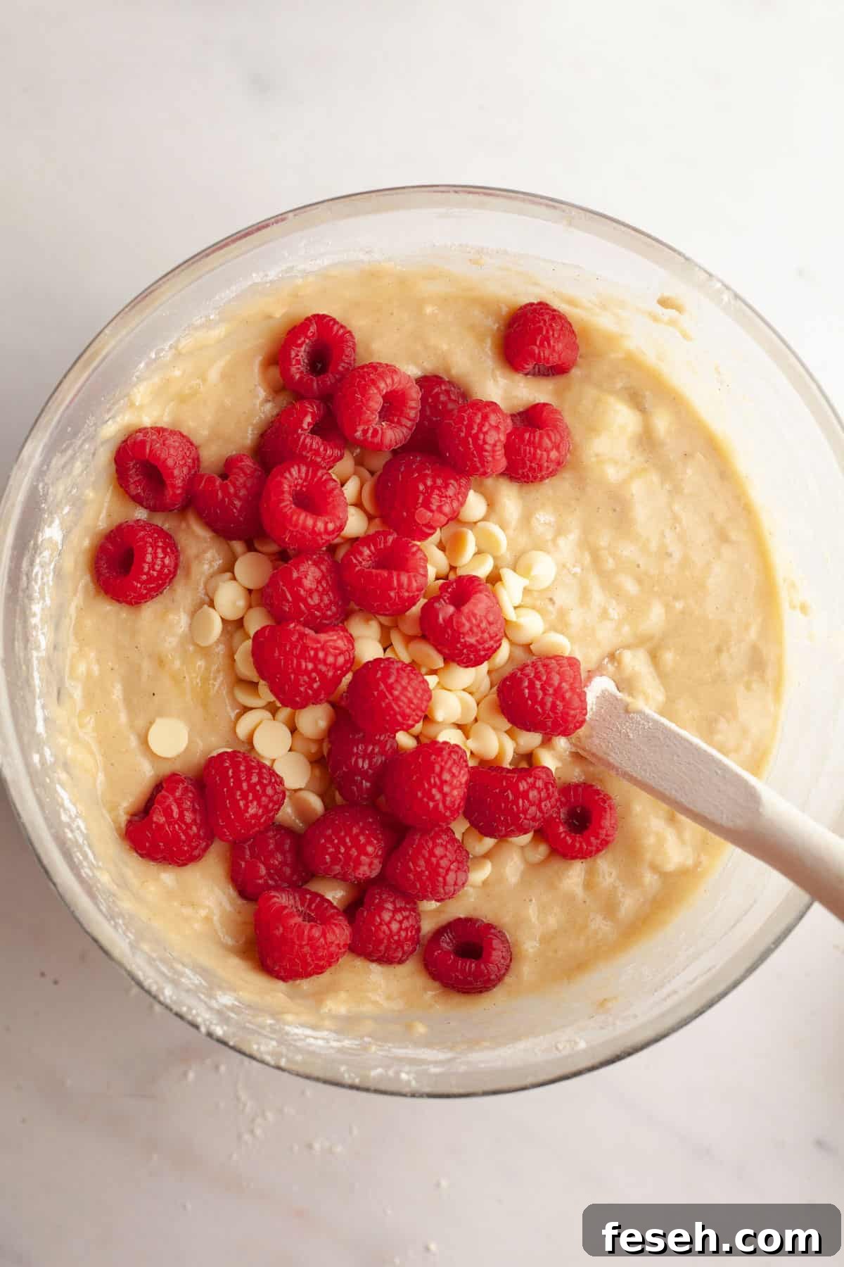 A large glass bowl with muffin batter in it with fresh raspberries and white chocolate chips.