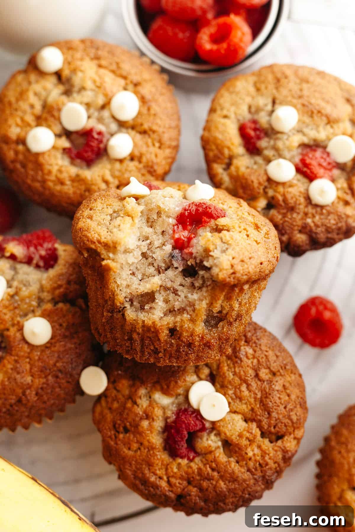 A close up of banana raspberry muffins with white chocolate chips, one has a bite taken out of it.