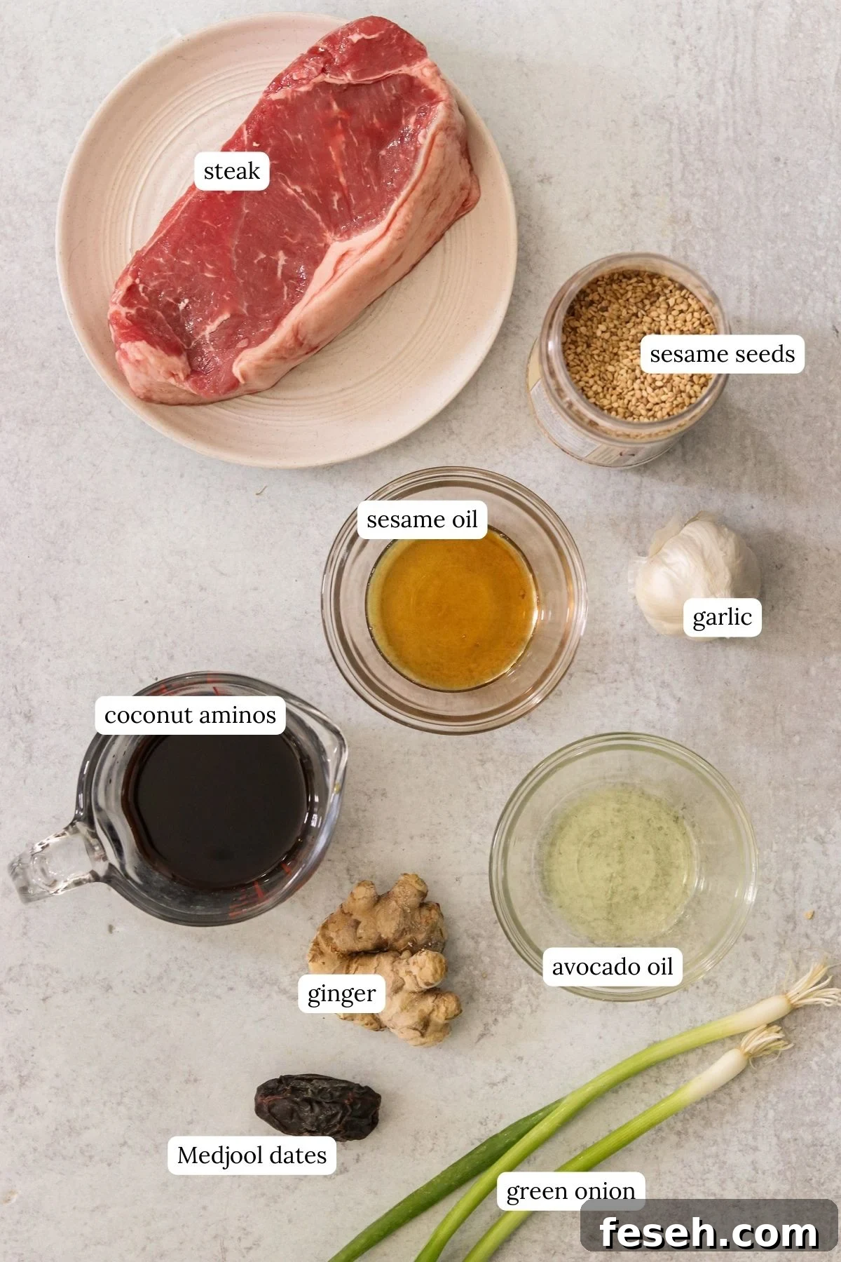 Overhead of labeled ingredients for Korean steak bites: sirloin, coconut aminos, sesame oil, garlic, ginger, date, gochugaru, oil, sesame seeds, and green onions.