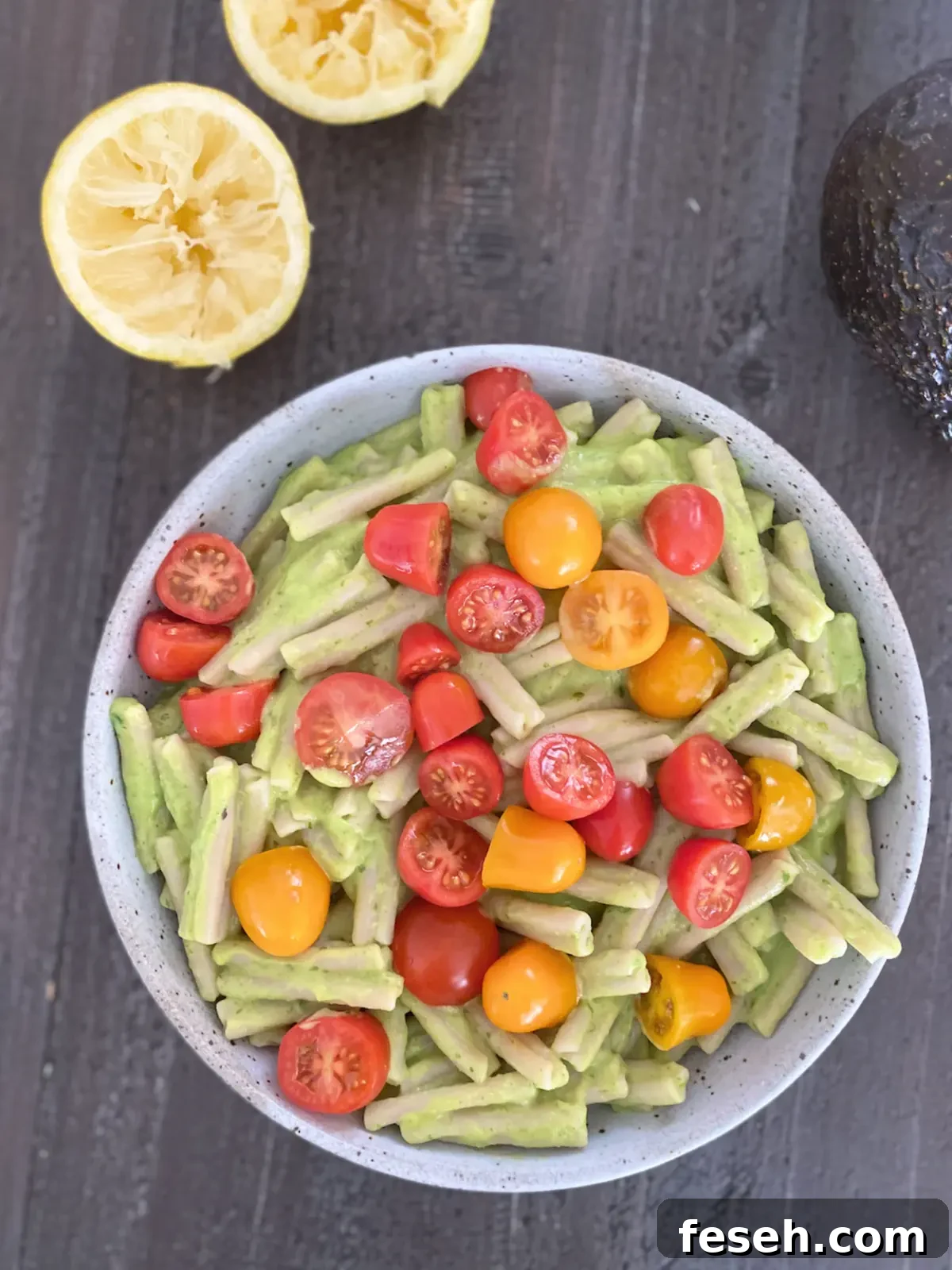 an overhead view of a bowl of pasta with light green avocado sauce and sliced tomatoes