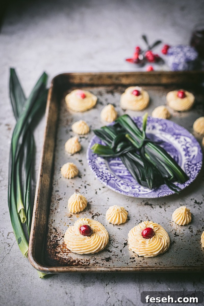 A baking sheet with piped Indonesian butter cookie dough in various sizes and in the middle of the sheet is a blue and white plate with tied bunches of pandan leaves