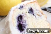 A slice of lemon blueberry bundt cake frosted with lemon cream cheese frosting sitting on a small white plate. Above the plate is a whole lemon and a blueberry lemon bundt cake frosted with lemon cream cheese frosting with a slice taken out of it.