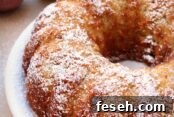 A Jewish Apple Bundt Cake dusted with powdered sugar on a white plate. In the upper left corner is a green and red apple. In the upper right corner is a powdered sugar shaker.