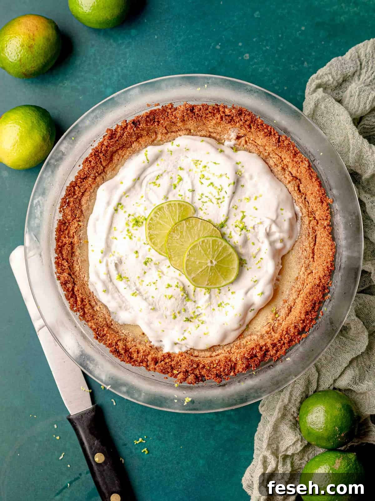 Key lime pie topped with whipped cream and lime slices next to limes on a table.