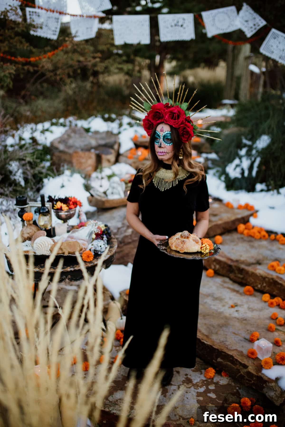 Woman dressed as a Catrina with face paint and a floral crown holding Pan de Muerto bread, standing beside a Día de los Muertos altar decorated with marigolds, papel picado, and pan dulce.