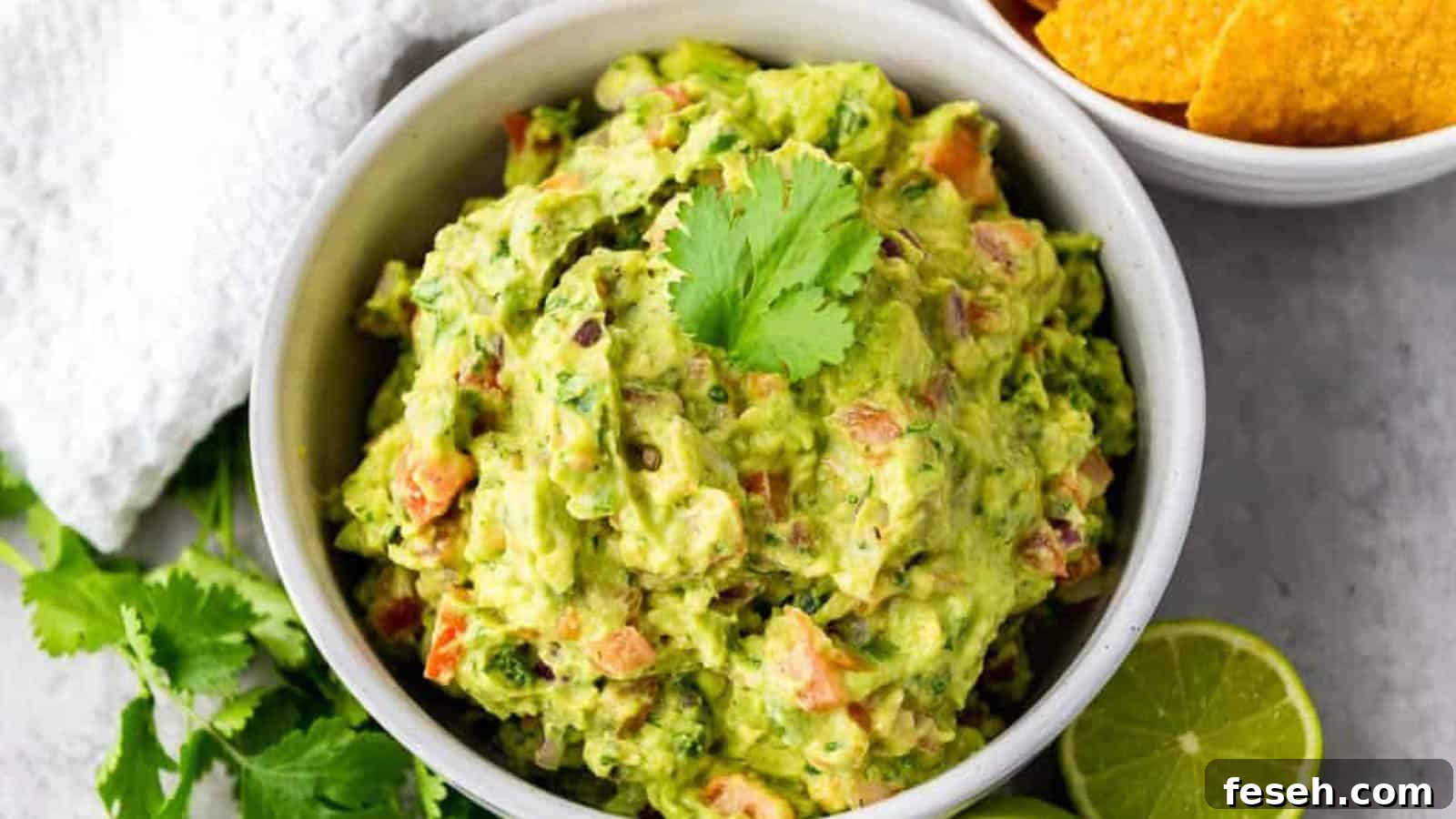 Guacamole in a white bowl with limes, cilantro, and tortilla chips on the side.