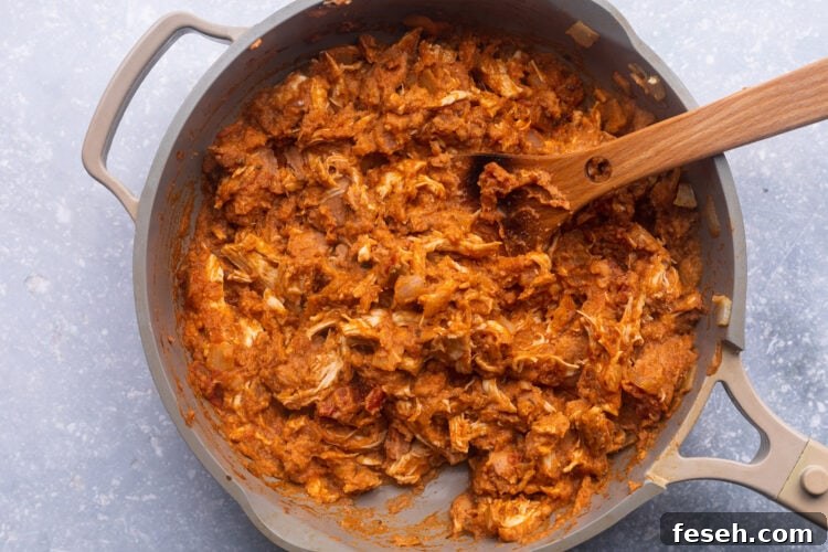 Shredded chicken, refried beans, salsa, taco seasoning, and onions in a large skillet with a wooden spoon.