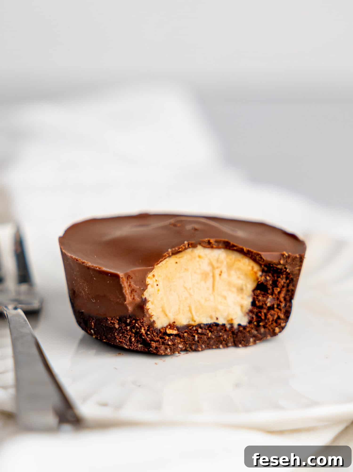 A peanut butter cup cheesecake on a small white plate, a cake fork to the left. A bite i missing from the cheesecake reavealing the creamy filling.