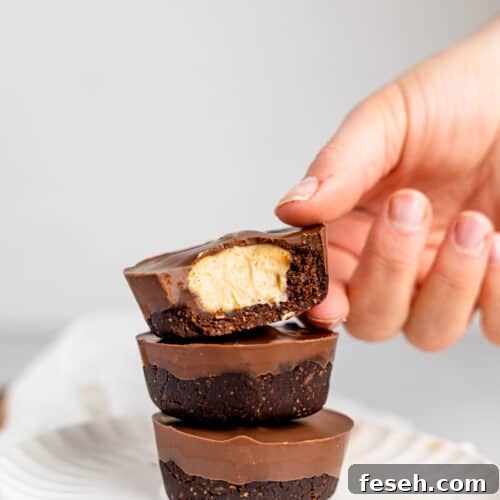 Three peanut butter cup cheesecakes stacked on top of each other on a white plate. A hand is slightly holding the top one and a bite is missing reavealing the cheesecake filling.