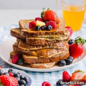 A stack of high protein french toast waffles sits on a white place on a table. Fresh berries and a slice of butter sit on top of the stack with maple syrup drizzling from a cup.