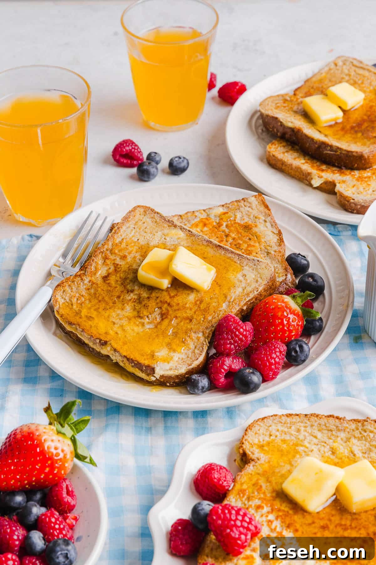 Two slices of french toast on a plate with butter and maple syrup on top. Fresh raspberries and blueberries are on the plate to the side for serving.