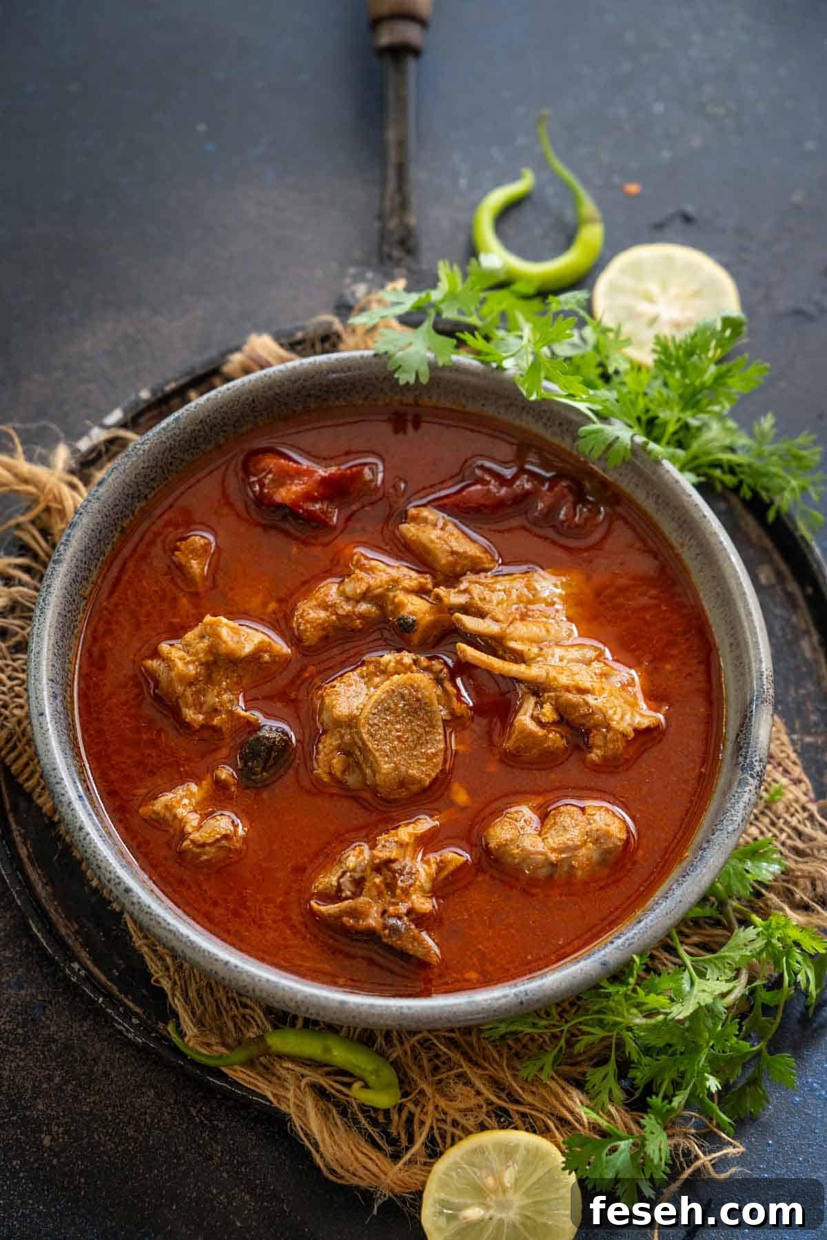 A bowl of spicy red Lamb Rogan Josh with tender pieces of meat, garnished with fresh cilantro, whole green chilies, and lemon slices on the side, served on a rustic dark surface.