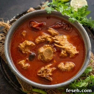 A bowl of spicy red Lamb Rogan Josh with tender pieces of meat, garnished with fresh cilantro, whole green chilies, and lemon slices on the side, served on a rustic dark surface.