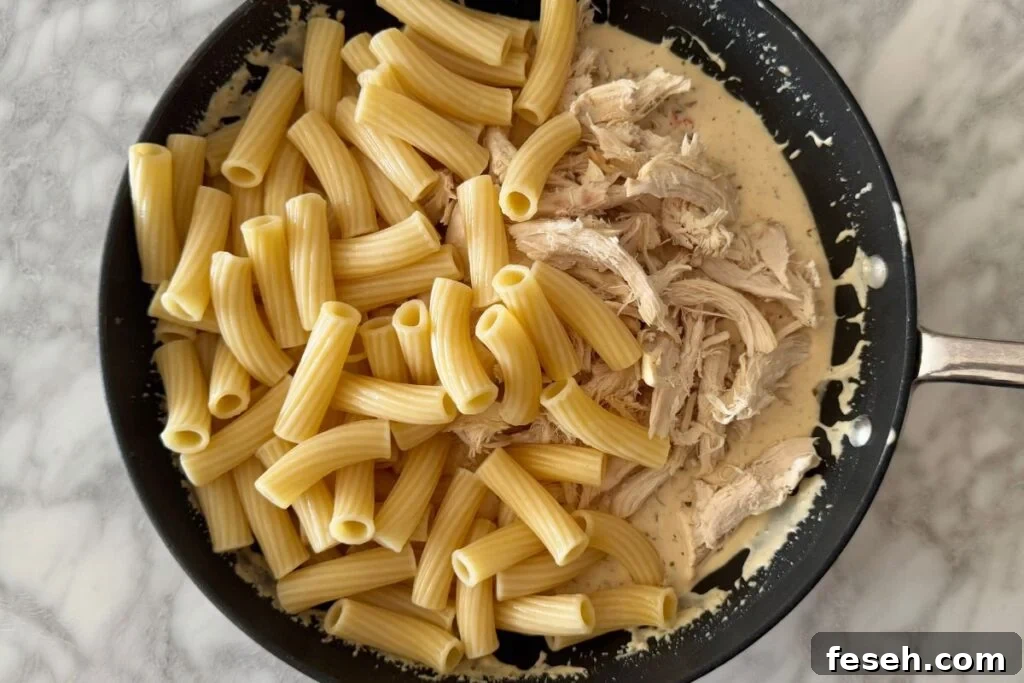 Shredded chicken and cooked pasta in skillet with sauce.