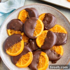 A mottled blue plater of candied orange slices dipped in chocolate with oranges and kitchen equipment around them.