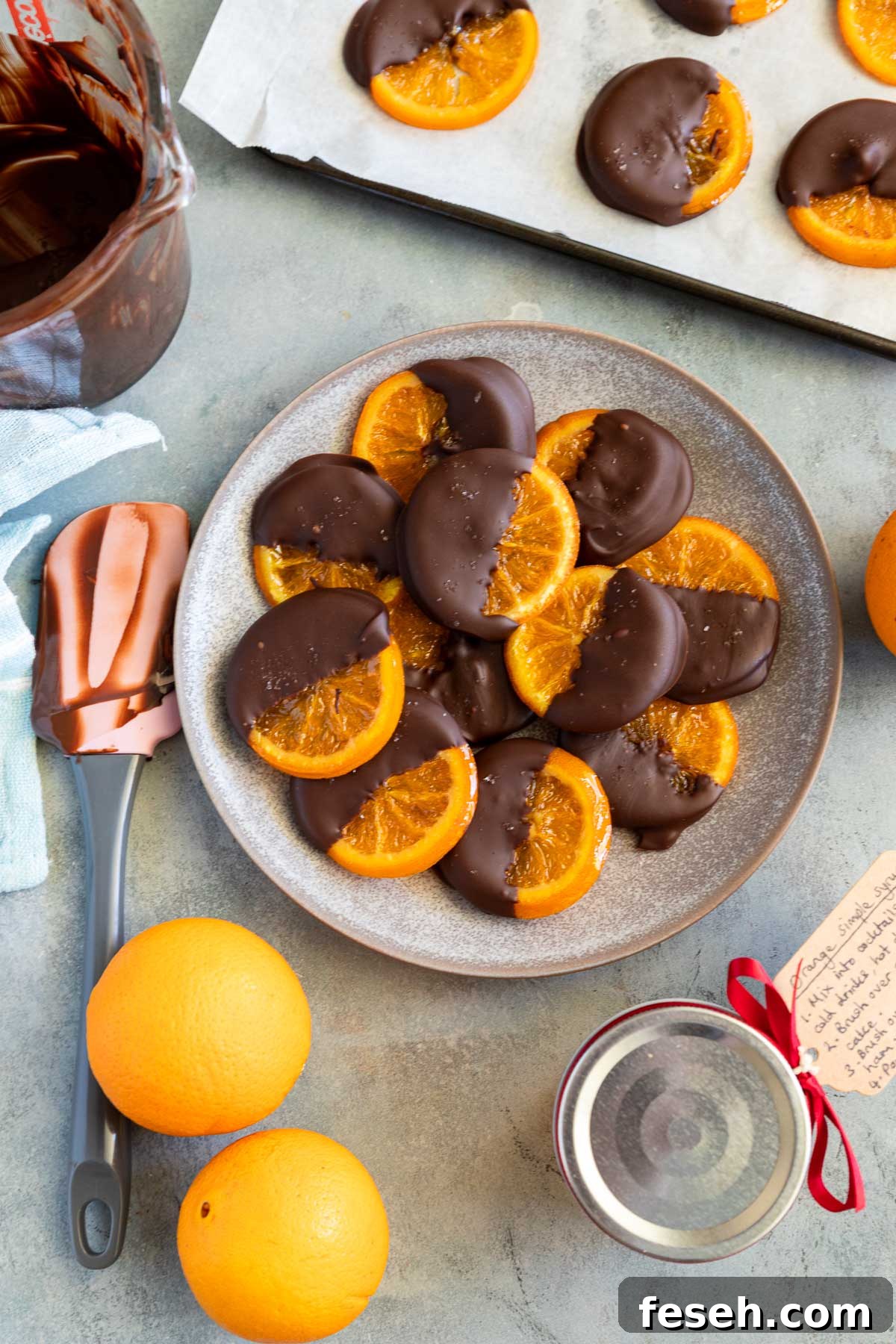 A mottled blue plater of candied orange slices dipped in chocolate with oranges and kitchen equipment around them.