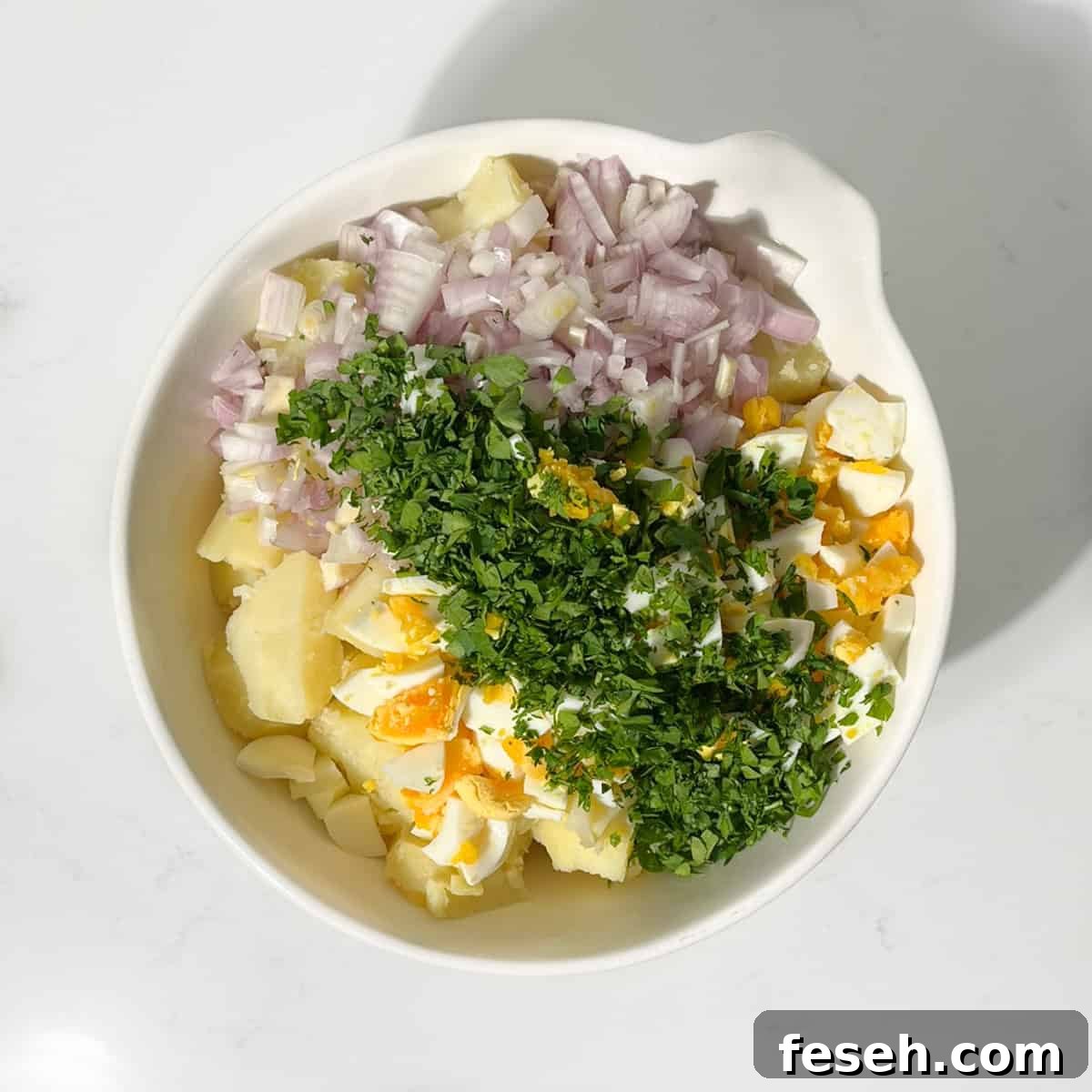 Adding the herbs, boiled eggs and onion to the sliced potato in a large white bowl.