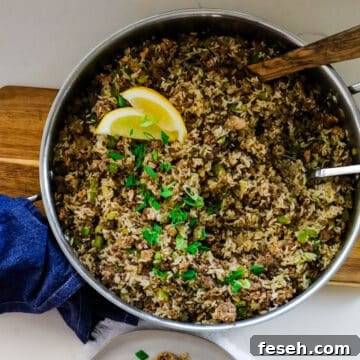 dirty rice in a large pan with a wooden spoon and green onions and lemon wedges as garnish.