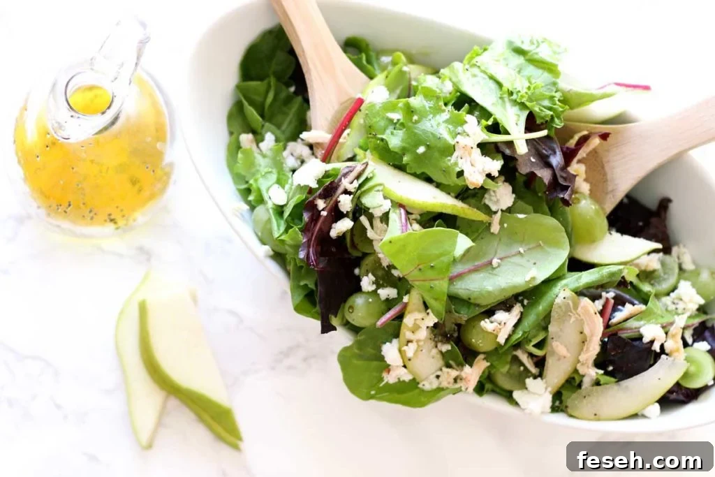 White serving dish containing a Pear and Goat Cheese Bistro Salad with two wooden serving spoons, mango dressing and sliced pear on white marble table.