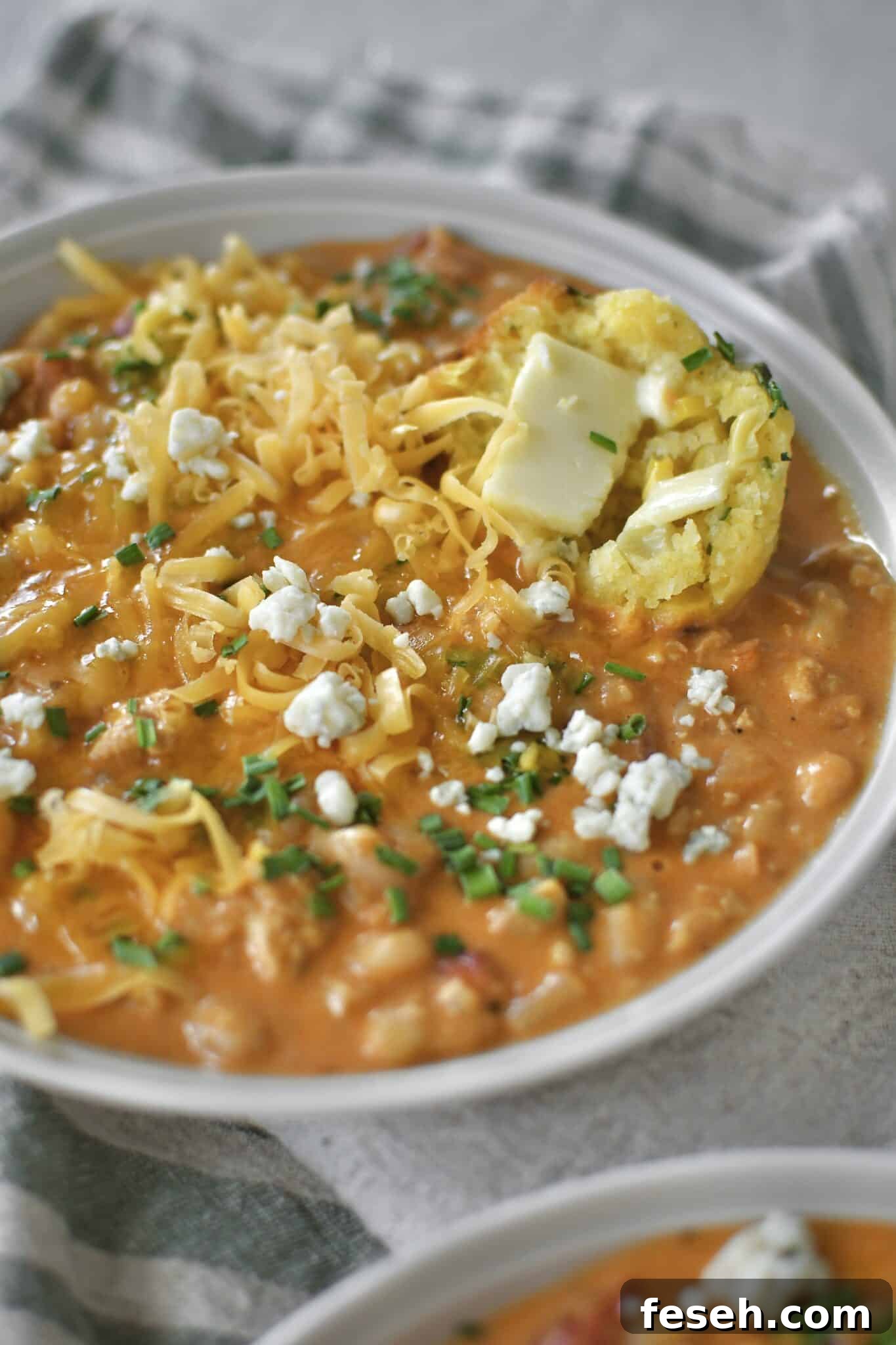 Buffalo Chicken Chili served in a bowl, topped with chives, blue cheese, and cheddar cheese. And a piece of cornbread muffin dunked in it.